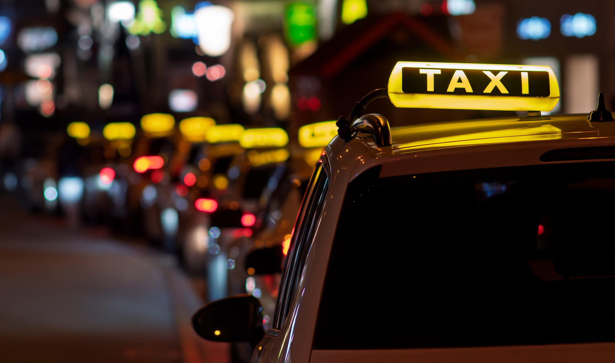 A row of taxis with illuminated signs wait in traffic on a city street at night, with blurred colorful lights in the background.