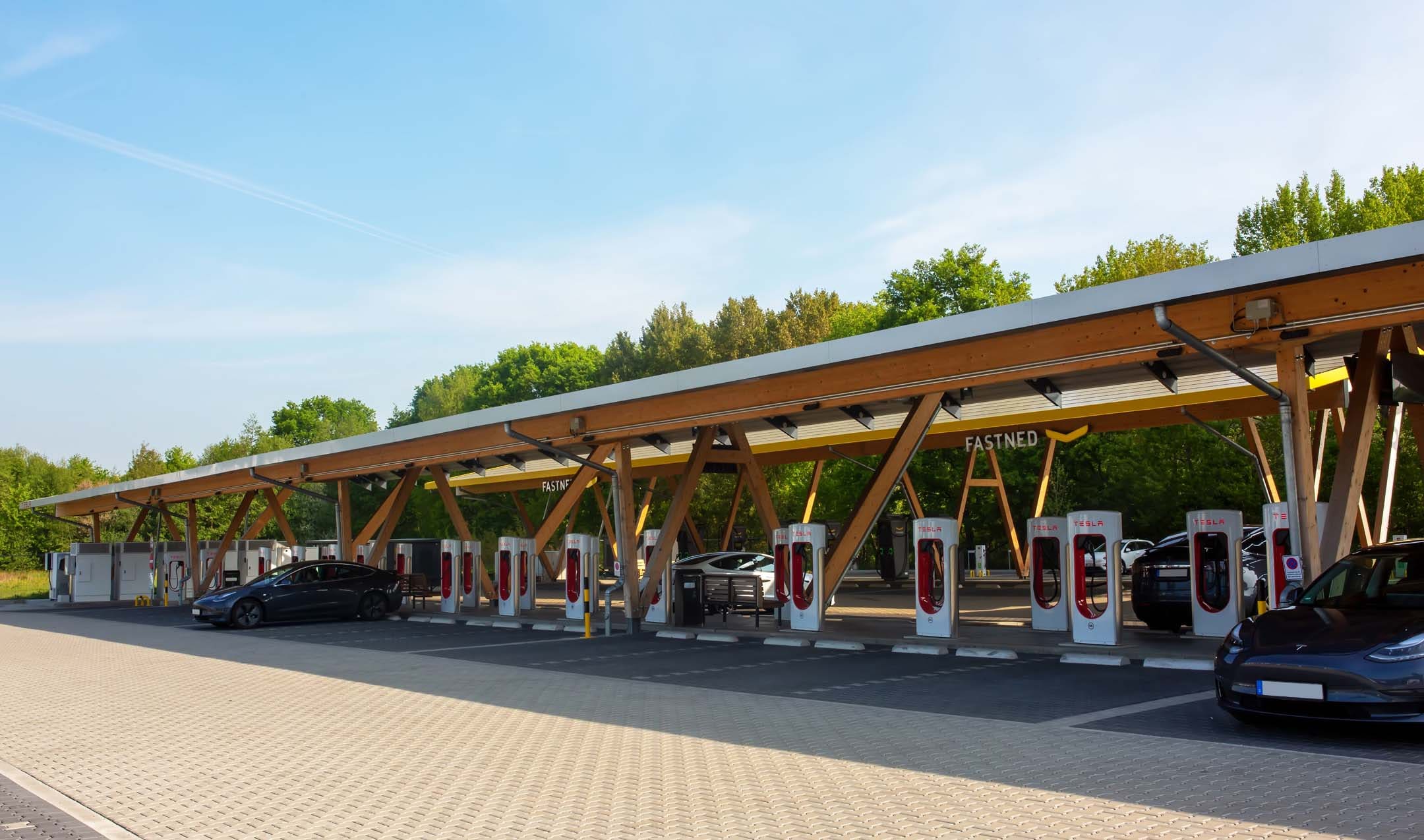 EV charging station under a wooden canopy with multiple Tesla Superchargers and parked cars, surrounded by greenery on a clear day.