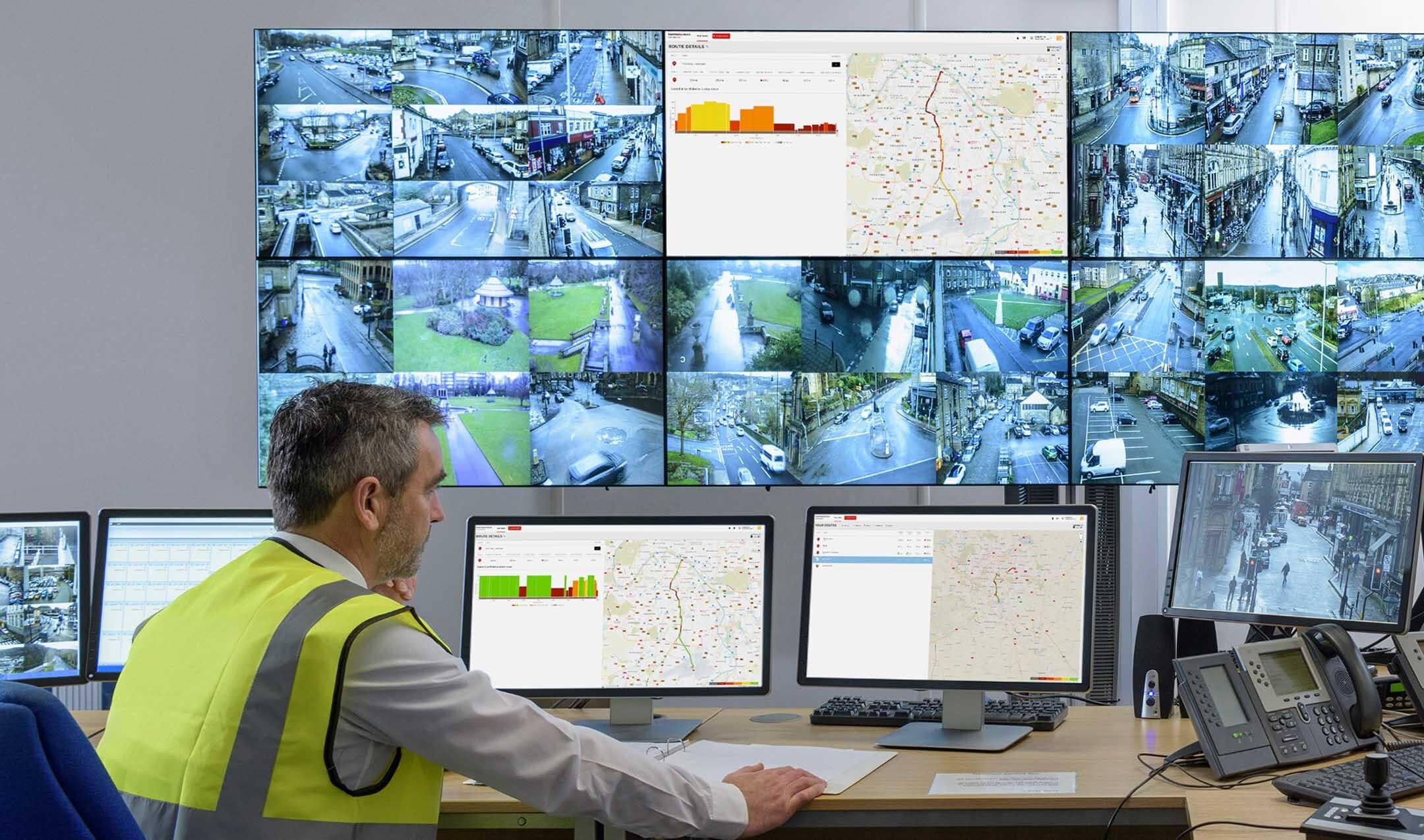 A man in a high-visibility vest monitors multiple CCTV and traffic management screens in a control room.