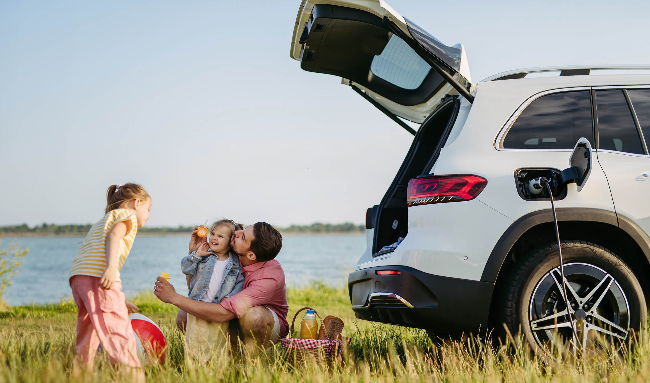 A family enjoys a picnic by a lake with a white electric SUV charging nearby; a father kisses a child while another plays with a toy.