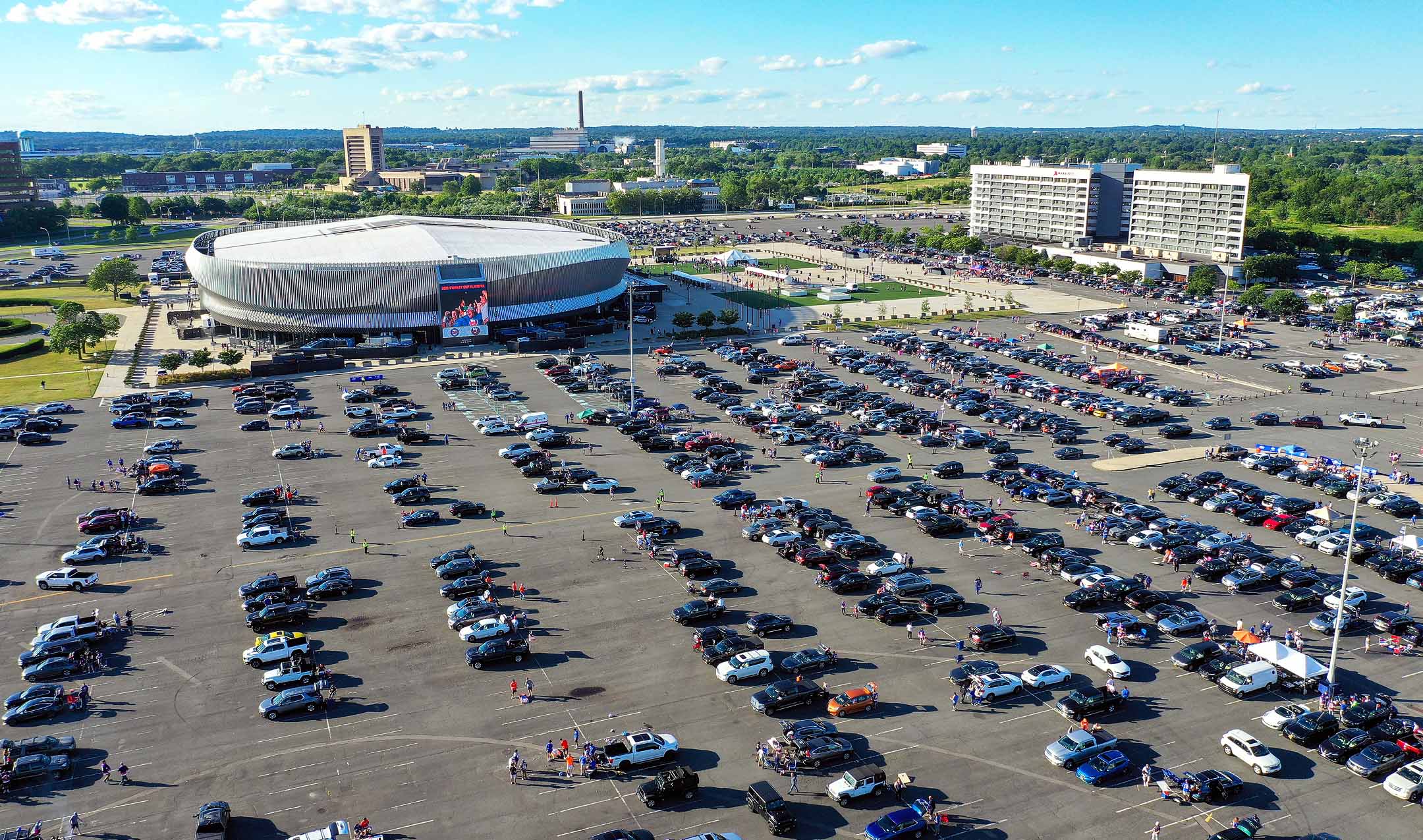 Aerial view of a large parking lot filled with cars outside a dome-shaped arena, with surrounding greenery and buildings under a clear blue sky.