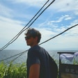 Tom in sunglasses stands by the ocean under a blue sky, with greenery and a sign in the foreground.