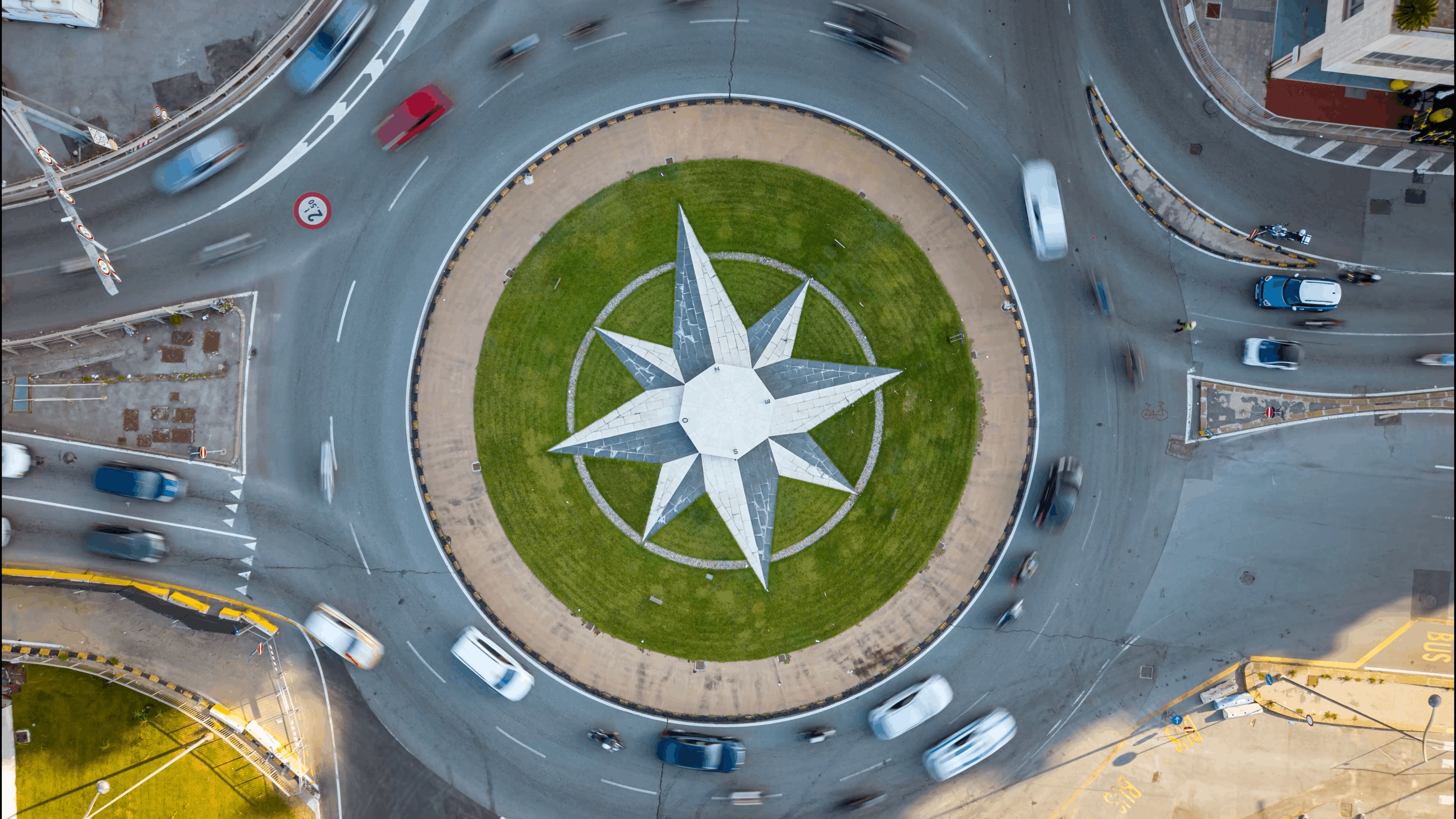 Aerial view of a roundabout with a large compass design in the center, surrounded by moving vehicles on the road.
