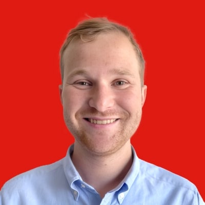 Smiling person with short light hair, wearing a light blue shirt, against a bright red background.